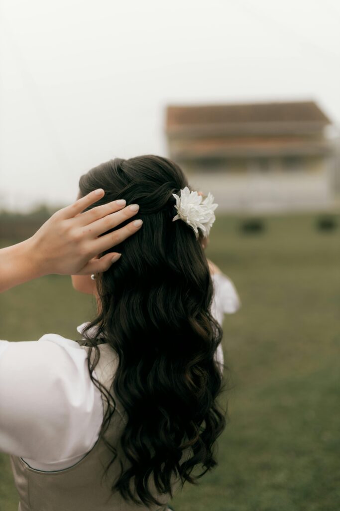 Novia de espaldas con melena suelta y ondas al agua marcadas, adornada con una flor blanca lateral, caminando en un jardín. Novia de espaldas con melena suelta y ondas al agua marcadas, adornada con una flor blanca lateral, caminando en un jardín.