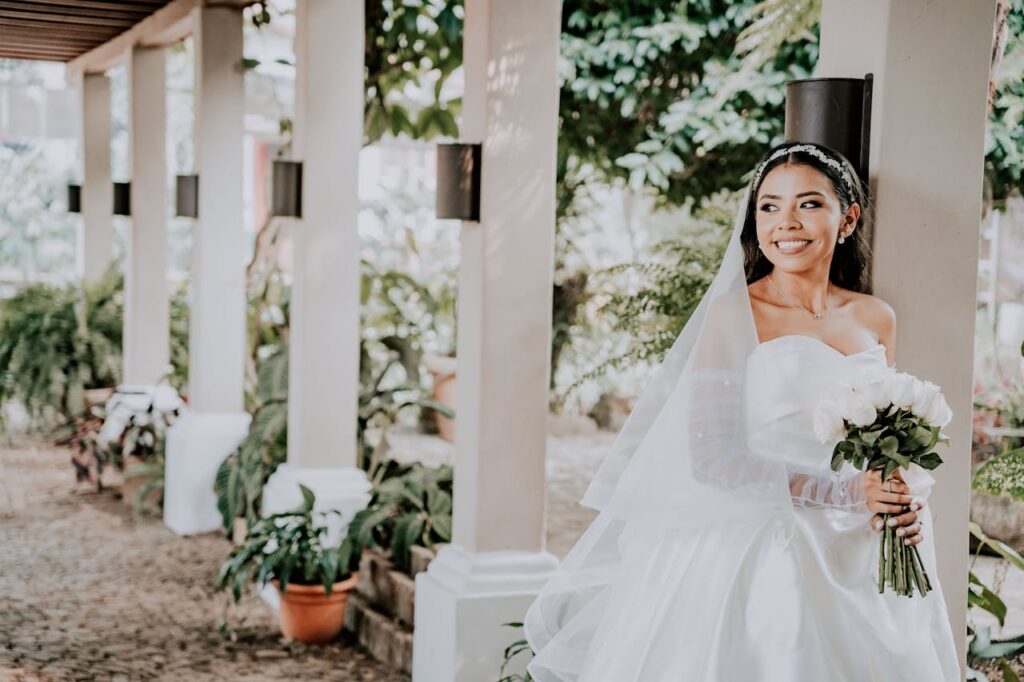 Novia sonriente con vestido de encaje y velo largo colocado sobre un peinado con tiara, posando en un porche con columnas blancas. Novia sonriente con vestido de encaje y velo largo colocado sobre un peinado con tiara, posando en un porche con columnas blancas.