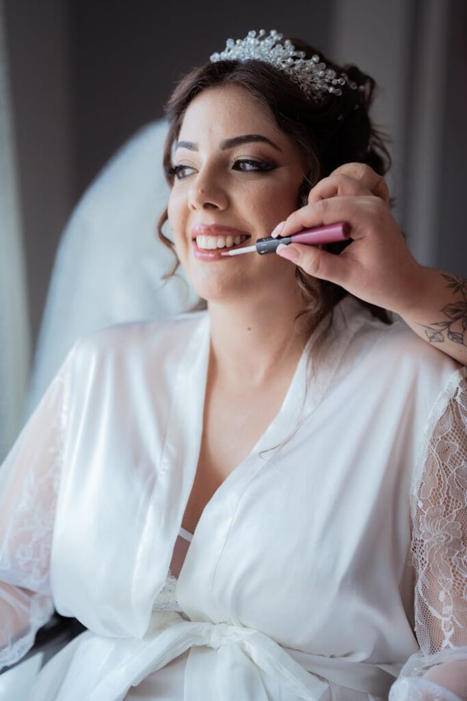 Primer plano de una novia maquillada con bata de satén blanca y sonriendo, sosteniendo una barra de labios rosa, representando la experiencia de Magaly Maquillaje en belleza nupcial a domicilio en Barcelona.